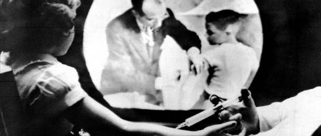 Une petite fille se fait vacciner contre la poliomyelite, le 12 avril 1955 a New York, tandis qu'elle regarde sur un ecran de television le Dr Jonas E. Salk, l'inventeur du vaccin, operer lui-meme a Ann Arbor, les vaccinations massives ayant commence sur tout le territoire des Etats-Unis.
